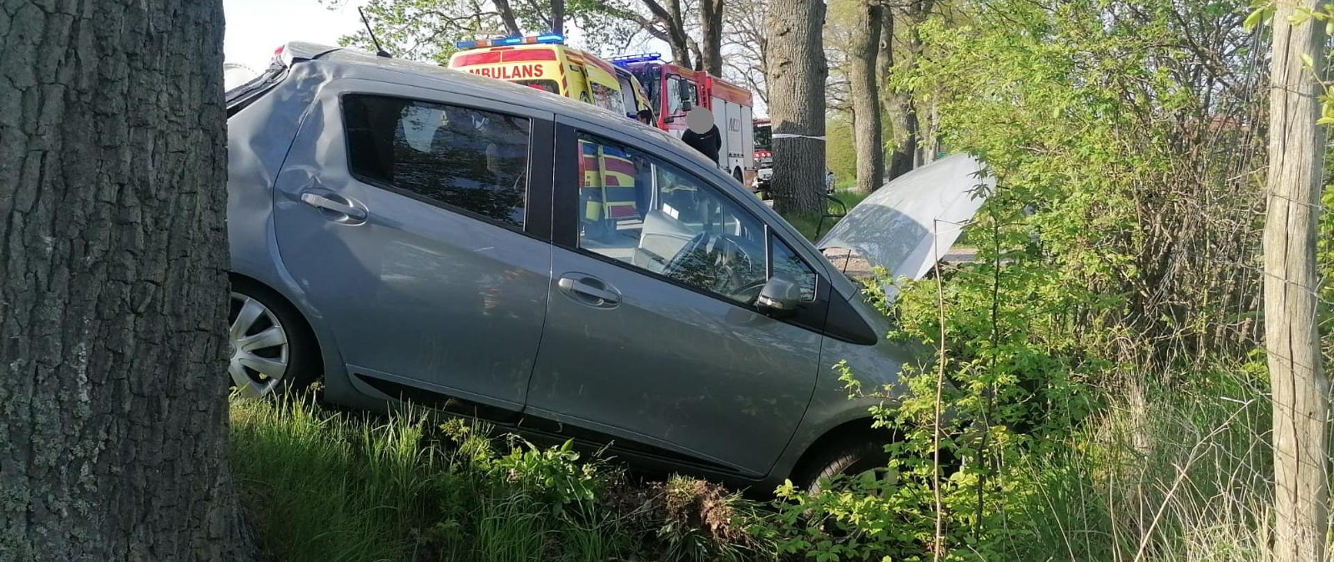 Szary samochód osobowy stoi w rowie. Dalej w oddali na drodze samochód strażacki i karetka. Z lewej i prawej strony drzewa.