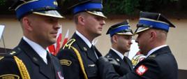 Zdjęcie zrobione na zewnątrz, podczas uroczystych obchodów Dnia Strażaka. Zdjęcie zrobione na placu przed jednostką ratowniczo-gaśniczą. Oficer Państwowej Straży Pożarnej w stopniu brygadiera przypina strażakowi medal.
