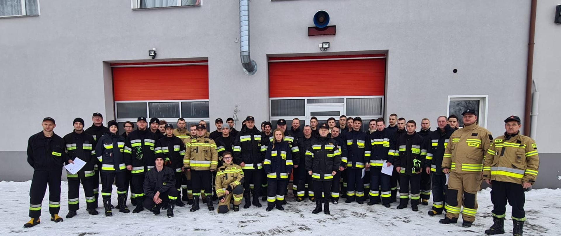 Zdjęcie wykonano za dnia na placu Komendy Powiatowej Państwowej Straży Pożarnej w Sępólnie Krajeńskim. Sceneria zimowa, na ziemi leży śnieg, kilkudziesięciu druhów Ochotniczych Straż Pożarnych pozuje do zdjęcia razem z Komendantem Powiatowym przed budynkiem komendy w ubraniach specjalnych. 