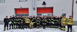 Zdjęcie wykonano za dnia na placu Komendy Powiatowej Państwowej Straży Pożarnej w Sępólnie Krajeńskim. Sceneria zimowa, na ziemi leży śnieg, kilkudziesięciu druhów Ochotniczych Straż Pożarnych pozuje do zdjęcia razem z Komendantem Powiatowym przed budynkiem komendy w ubraniach specjalnych. 