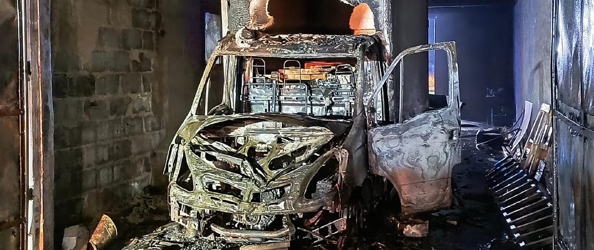 Na zdjęciu widać spalony samochód dostawczy, który stoi wewnątrz zniszczonego budynku, którego dach i konstrukcja stropowa są zwęglone. Wnętrze furgonetki jest całkowicie wypalone, a dookoła widać pozostałości po pożarze.