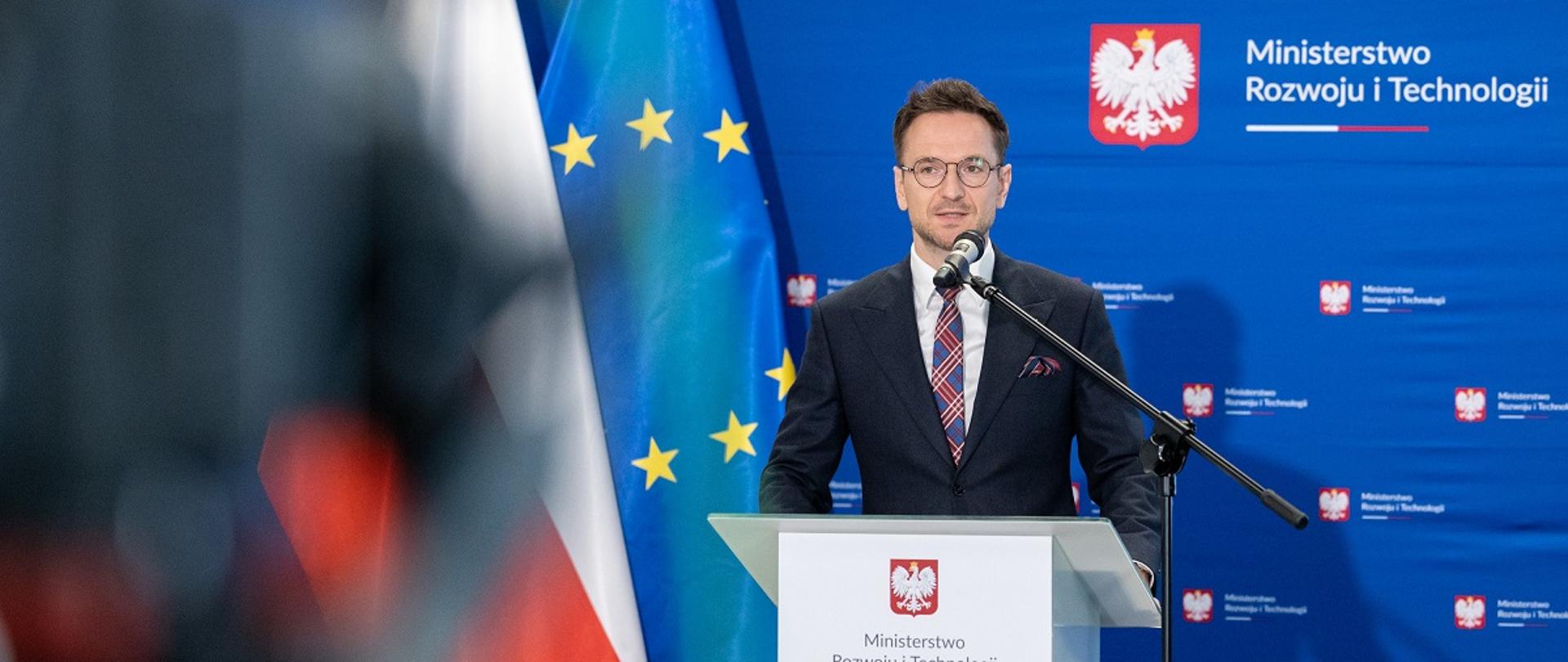 Minister rozwoju i technologii Waldemar Buda na konferencji prasowej poświęconej zmianom w przepisach dotyczących gwarantowanych kredytów mieszkaniowych, minister stoi przy mównicy z mikrofonem, za jego plecami flagi Polski i UE oraz baner MRiT