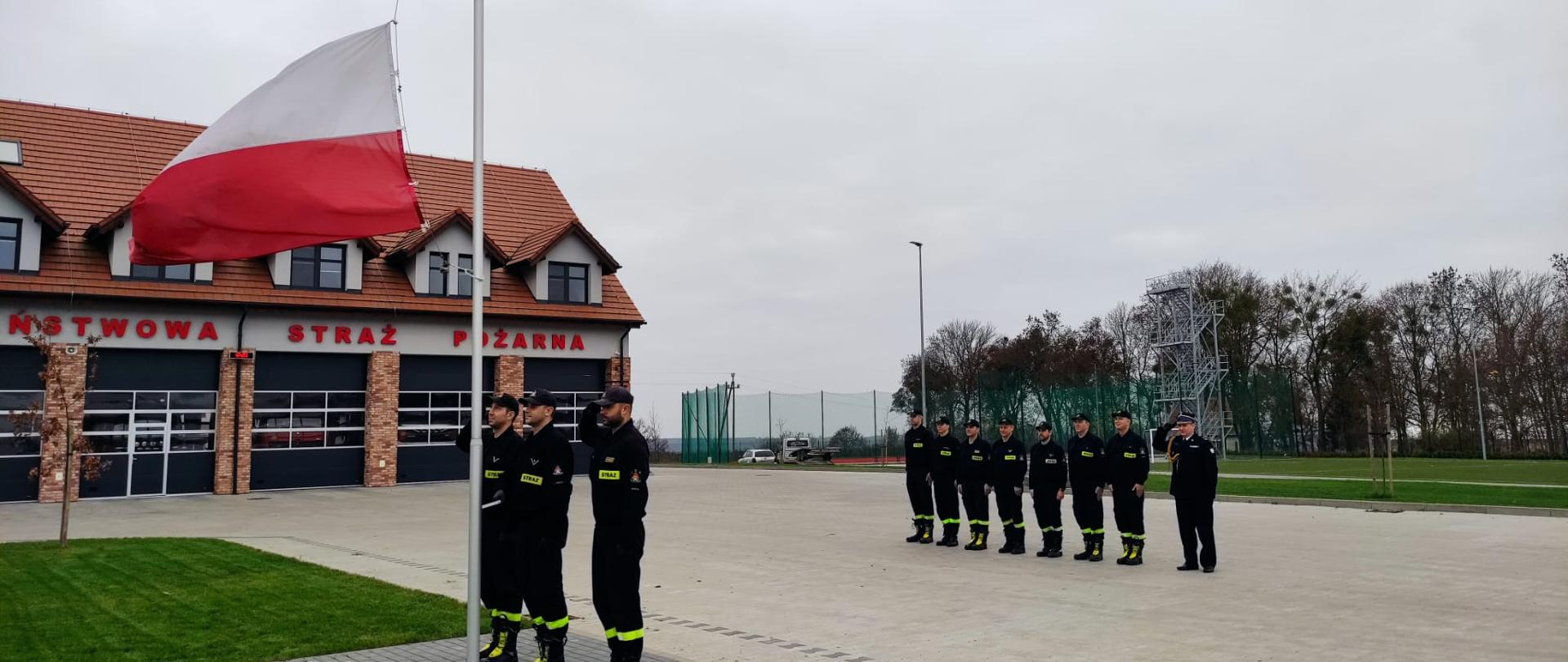 Na placu Komendy Powiatowej Państwowej Straży Pożarnej w Świeciu od lewej przed masztem na który jest wciągana flaga narodowa stoją trzej strażacy, za nimi w szeregu ośmiu funkcjonariuszy. 