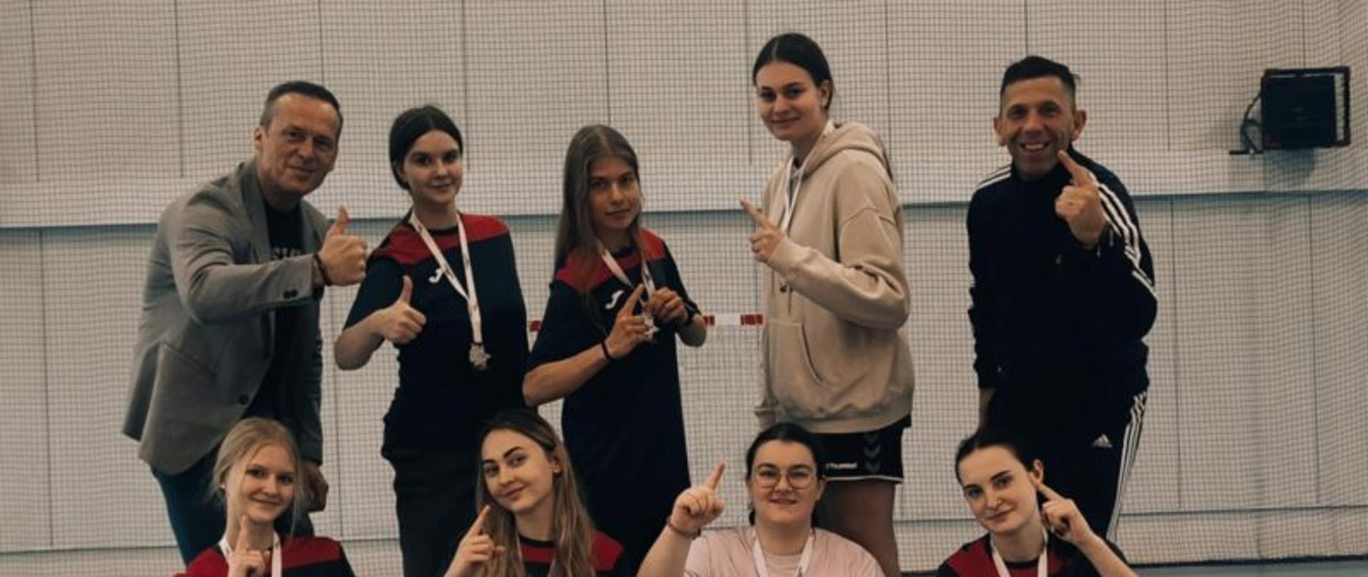 Grupa uczniów w strojach do koszykówki pozuje w sali gimnastycznej. Z uczniami stoją dyrektor i nauczyciel wychowania fizycznego.