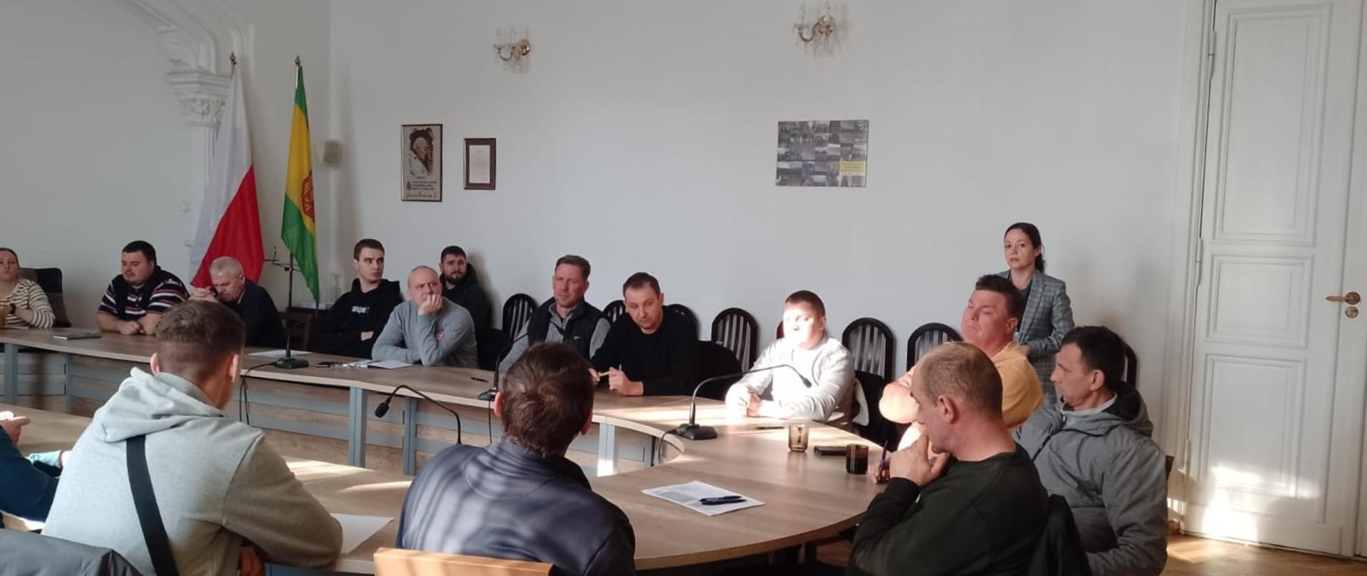 Sala, przy owalnym stole siedzą ludzie. Przy ścianie stoi kobieta w marynarce.