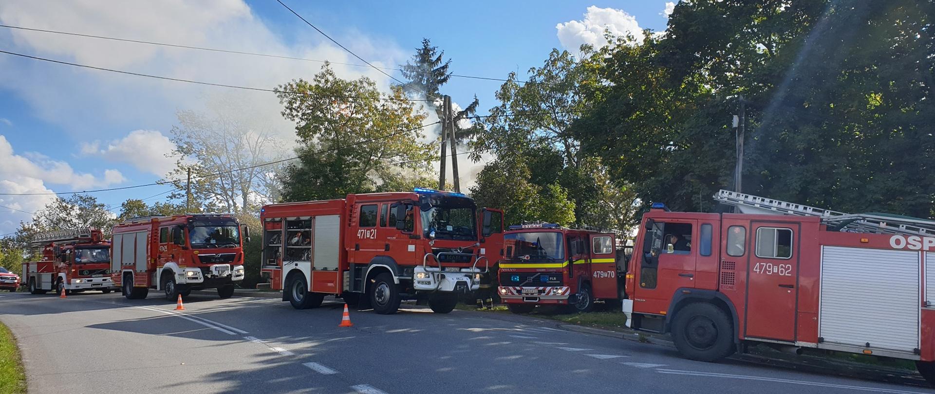 Na pierwszym planie widać samochody pożarnicze, w tle dym z palącego się budynku.