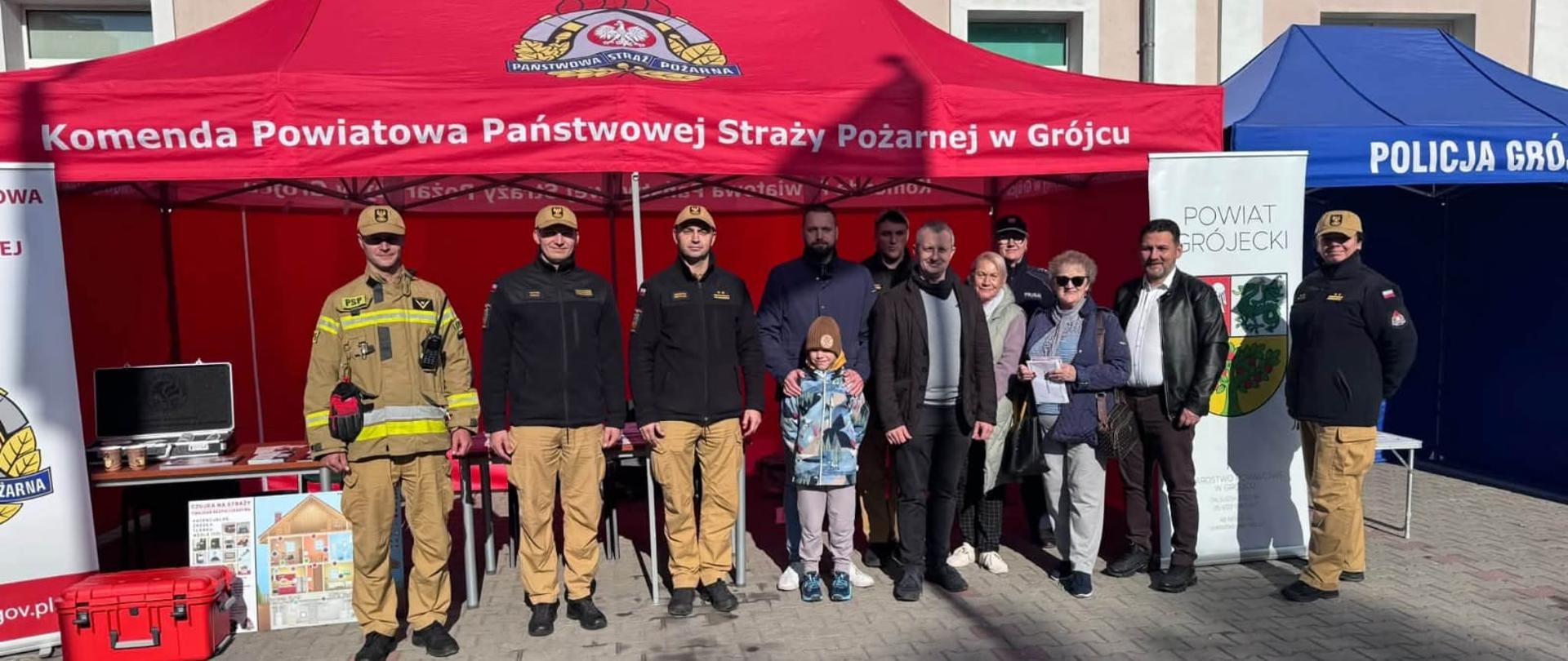 Przed czerwonym namiotem Komendy Powiatowej Państwowej Straży Pożarnej w Grójcu stoi grupa strażaków, przedstawicieli lokalnej społeczności i odwiedzających, pozujących do wspólnego zdjęcia. Strażacy mają na sobie czarne mundury z beżowymi spodniami lub jasne ubrania specjalne z odblaskowymi pasami. Wśród pozostałych osób znajduje się dziecko w kolorowej kurtce. Pod namiotem ustawiono stoły z materiałami informacyjnymi i sprzętem. Po lewej stronie widoczna jest plansza edukacyjna dotycząca bezpieczeństwa w domu oraz czerwona skrzynia ratownicza. Po prawej stronie znajduje się stojak z herbem powiatu grójeckiego, a w tle niebieski namiot Policji. Wydarzenie odbywa się na nasłonecznionym placu przed budynkiem.