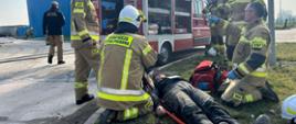 Na pierwszym planie punkt udzielania kwalifikowanej pierwszej pomocy. Na tle lekkiego samochodu pożarniczego, na noszach leży poszkodowany pracownik. W głębi dwa zbiorniki. Przy zbiorniku z lewej strony znajduje się 2 strażaków prowadzących działania gaśnicze. 