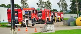Samochody pożarnicze ustawione jeden za drugim podczas ćwiczeń. Przy samochodach znajdują się strażacy. 