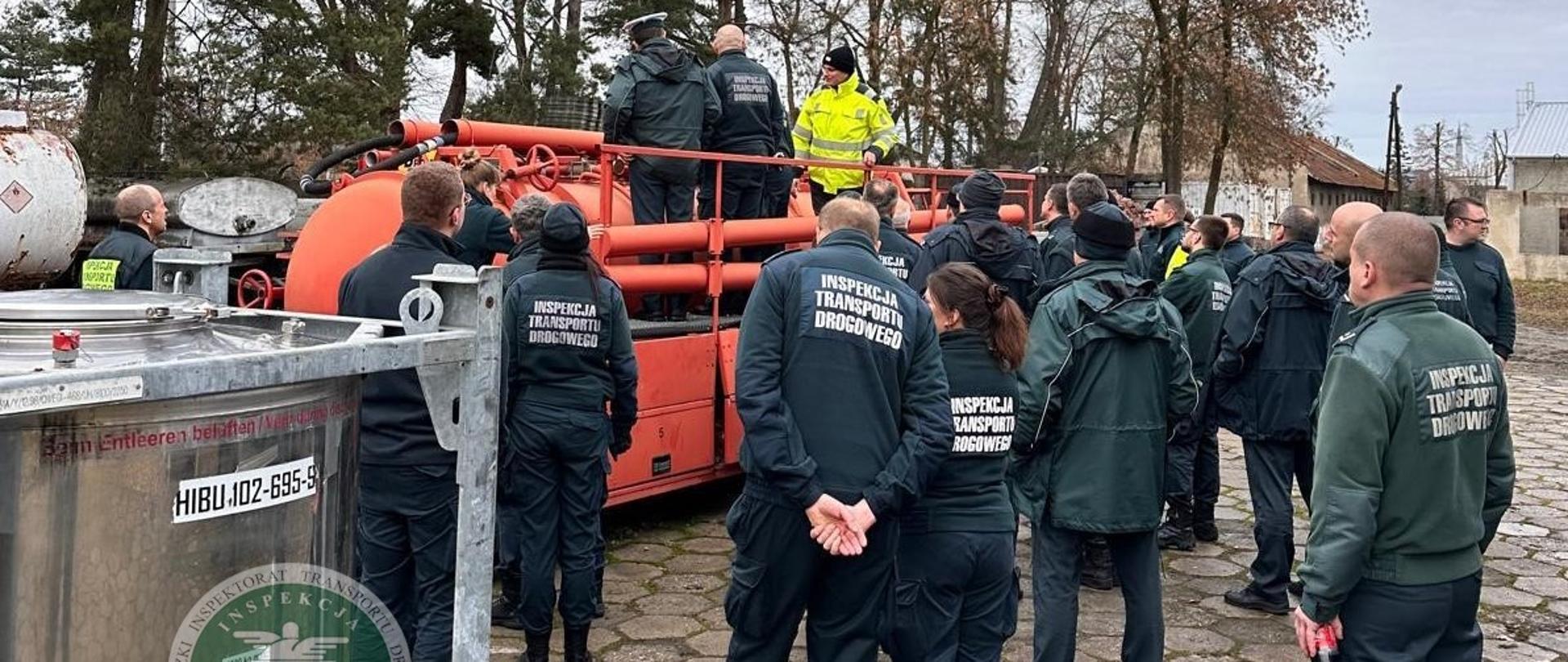 Pochmurny dzień, grupa około 20 osób w zielonych mundurach z napisem „INSPEKCJA TRANSPORTU DROGOWEGO” stoi zwrócona do pomarańczowej cysterny znajdującej się na betonowych płytach. Dwie osoby oraz jeden mężczyzna w jaskrawej, żółtej bluzie stoją na cysternie przyglądając się jej konstrukcji. W tle widać kilka budynków gospodarczych oraz drzewa. 