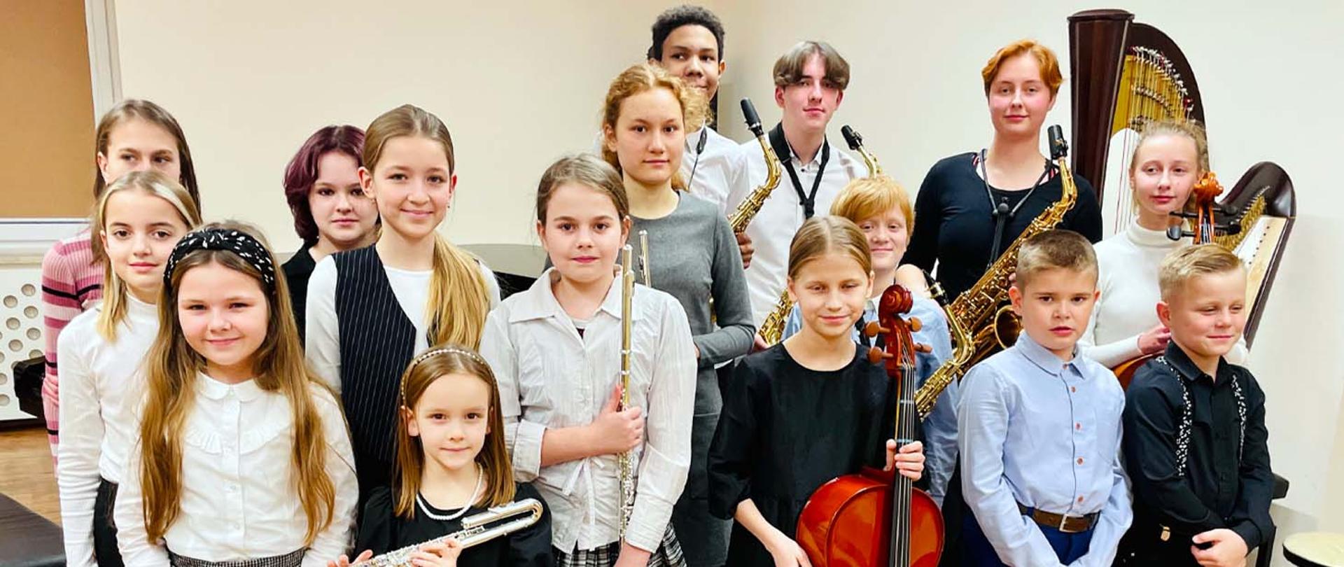 Na zdjęciu, w pomieszczeniu z jasnymi ścianami stoi grupa kilkunastu uśmiechniętych dzieci, patrzą przed siebie, niektórzy z nich w rękach trzymają instrumenty muzyczne. Za nimi po prawej stronie stoi harfa a po środku fortepian.