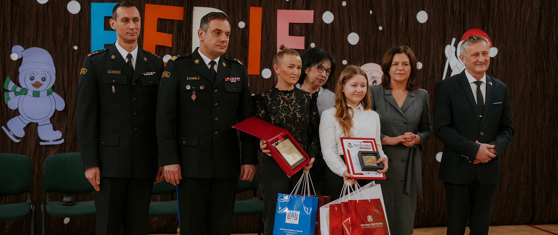 Pamiątkowe zdjęcie grupowe. Od lewej: Komendant Miejski PSP w Kielcach bryg. Michał Jankowski, Świętokrzyski Komendant Wojewódzki Państwowej Straży Pożarnej nadbryg. Krzysztof Ciosek, mama odznaczonej dziewczynki Ewelina Góra, dyrektor szkoły podstawowej nr 24 pani Ewa Misiorowska, odznaczona odznaką Iuvenis Forti: Kinga Góra, świętokrzyski wicekurator oświaty pani Katarzyna Nowacka oraz Wojewoda Świętokrzyski Zbigniew Koniusz.