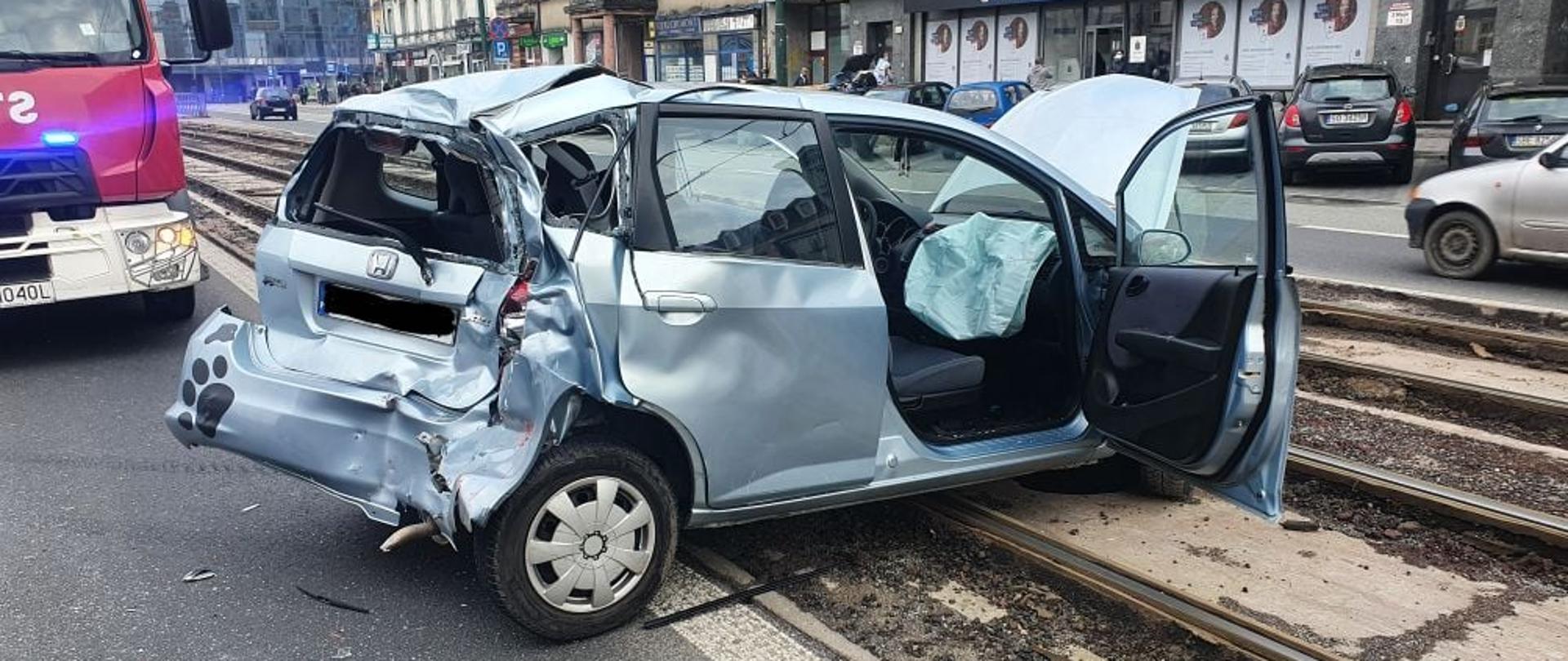Zdjęcie przestawia uszkodzony samochód koloru srebrnego znajdujący się na torach tramwajowych , w tle samochód straży pożarnej 