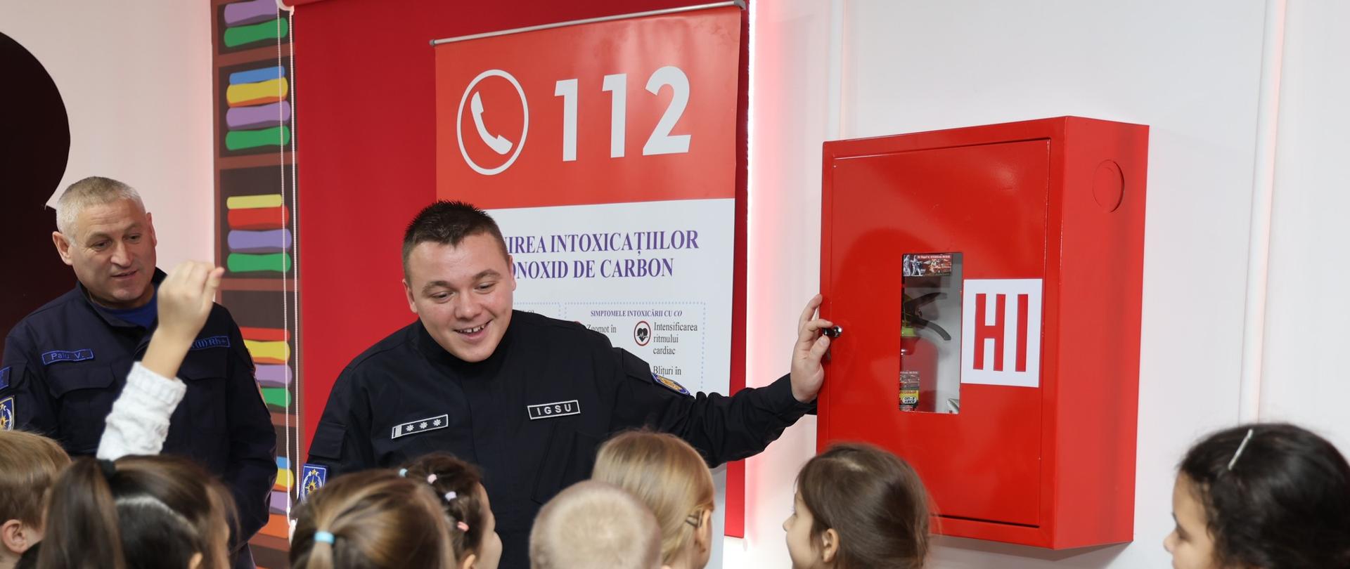 Strażak pokazuje dzieciom gaśnicę, grupka dzieci w wieku szkolnym go słucha, jedno dziecko podnosi rękę