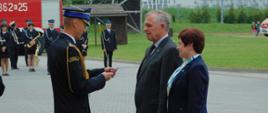 Na zdjęciu widoczny funkcjonariusz wręczający kobiecie i mężczyźnie medal. Funkcjonariusz ubrany jest w mundur galowy na głowie ma czapkę rogatywkę. Mężczyzna ubrany w garnitur, kobieta żakiet koloru granatowego. W tle widoczny samochód pożarniczy i orkiestra dęta.
