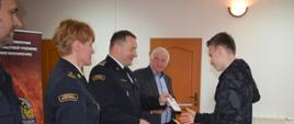 Komendant Powiatowy Państwowej Straży Pożarnej, ubrany w mundur, wręcza stojącemu na przeciw niego, chłopcu nagrodę za zajęcie miejsca w konkursie. Obok komendanta stoi Pani strażak, ubrana w mundur oraz przedstawiciel starostwa powiatowego, ubrany w garnitur.
