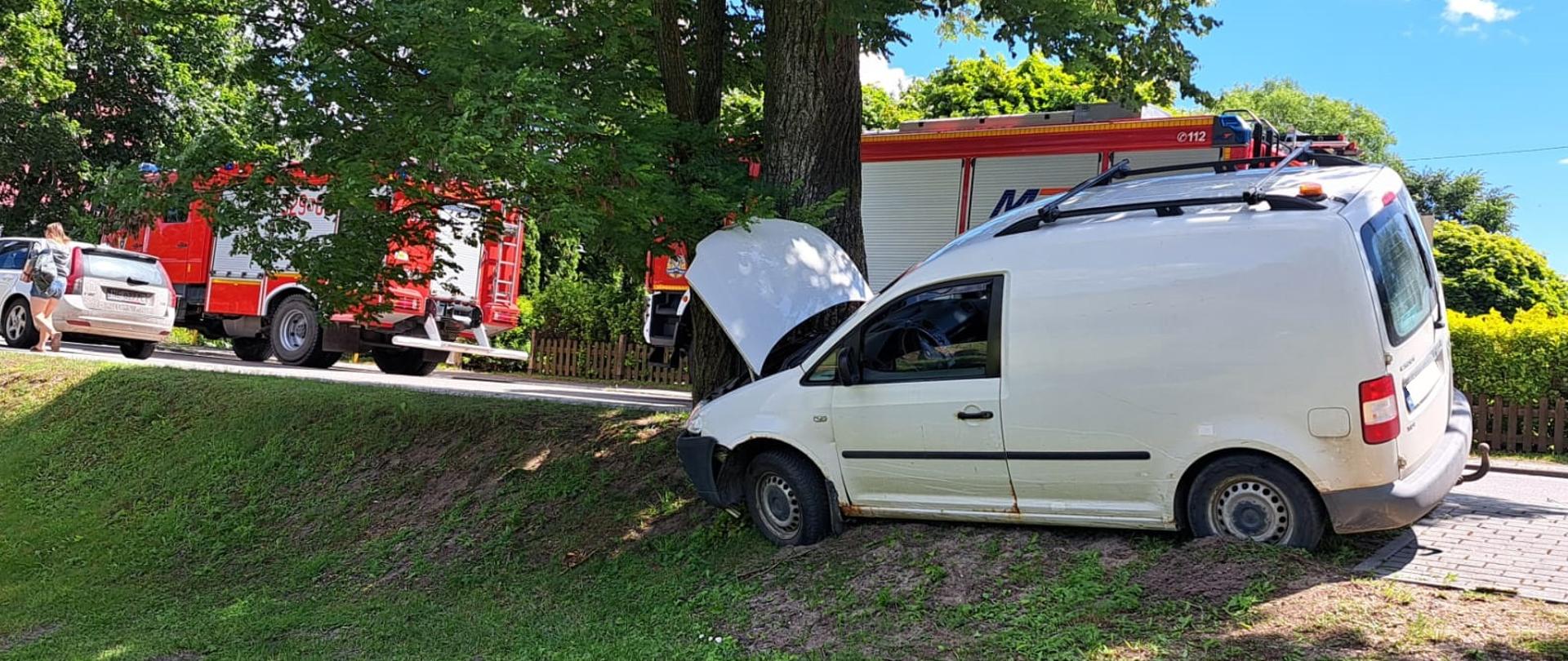 Pojazd osobowy VW Caddy koloru białego uderzył w przydrożne drzewo. W tle widoczne są 2 samochody pożarnicze oraz inny pojazd osobowy. 
