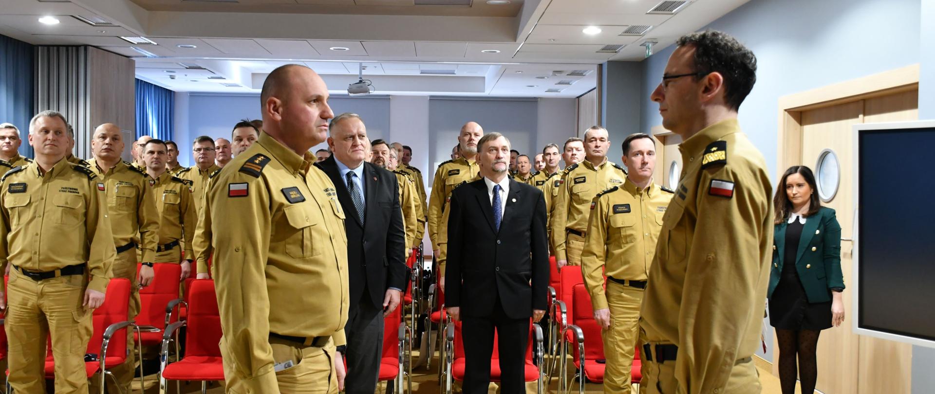 Uroczystość w sali konferencyjnej. Dwóch strażaków PSP w mundurach służbowych w kolorze żółtym stoi naprzeciw siebie. Za nimi zgromadzeni funkcjonariusze PSP oraz kilka osób w garniturach. Sala wyposażona w czerwone krzesła, jasne ściany i sufit z oświetleniem.