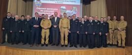 Zdjęcie grupowe, na scenie stoją zaproszeni goście ubrani w garnitury, oraz strażacy ubrani w mundury, z tyłu widać wyświetloną prezentację