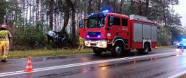 Samochód pożarniczy na drodze, obok strażak. W tle samochód pomiędzy drzewami zakleszczony