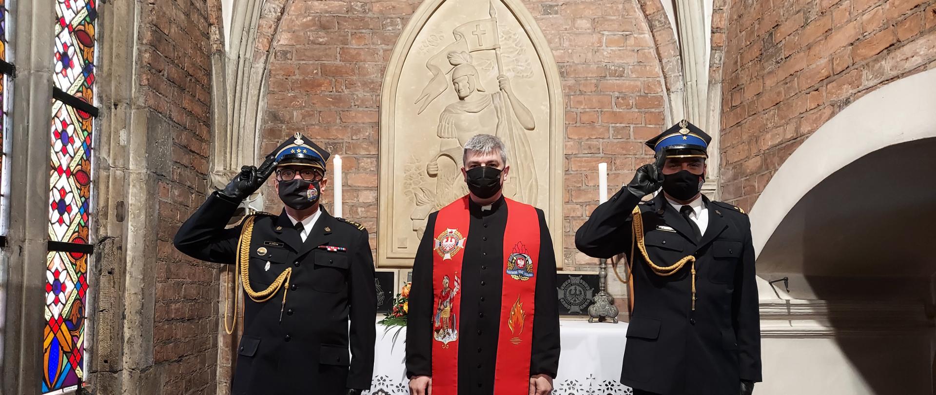 Strażacy i kapelan przed płaskorzeźbą św. Floriana