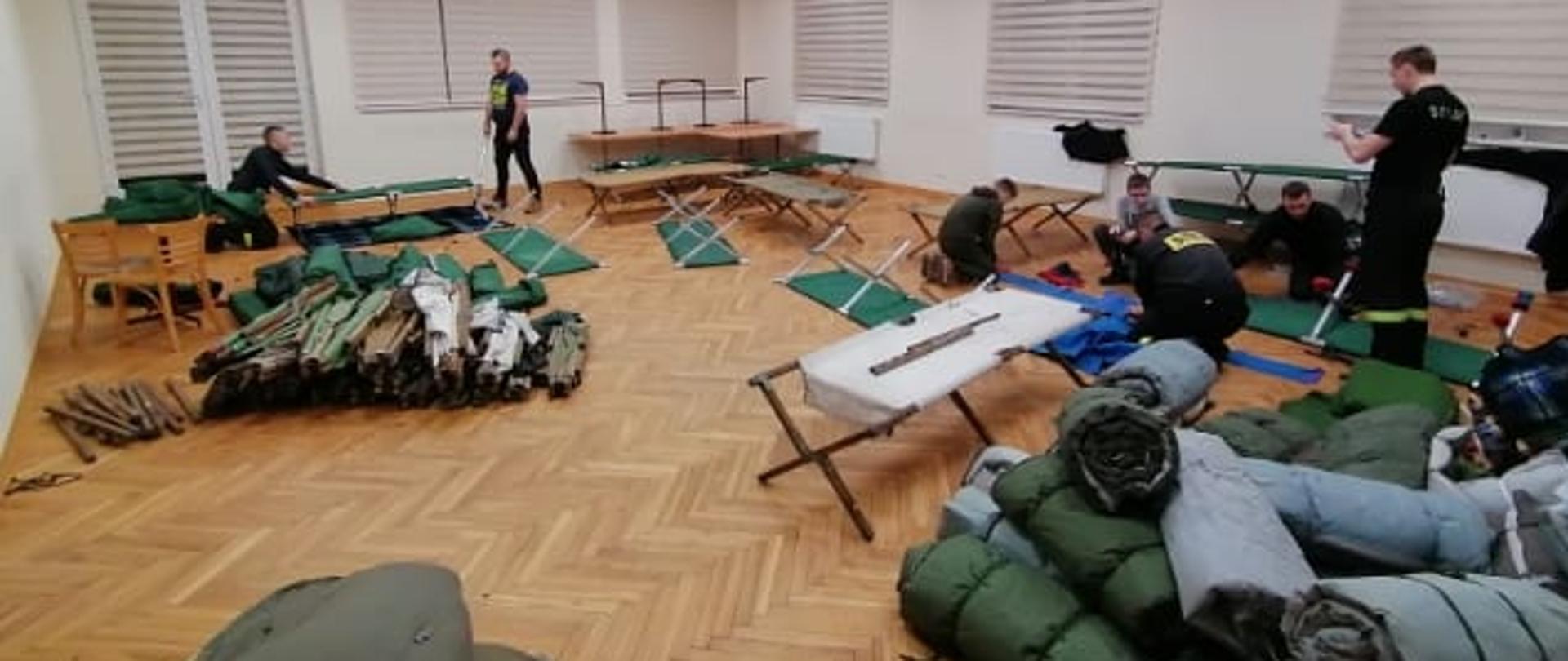 Na sali widocznych kilku strażaków. Po prawej stronie leżą zwinięte materace. Po lewej złożone łóżka polowe. W głębi sali dwie grupy strażaków rozkładają łóżka polowe.