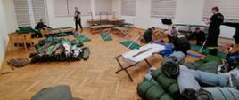 Na sali widocznych kilku strażaków. Po prawej stronie leżą zwinięte materace. Po lewej złożone łóżka polowe. W głębi sali dwie grupy strażaków rozkładają łóżka polowe.