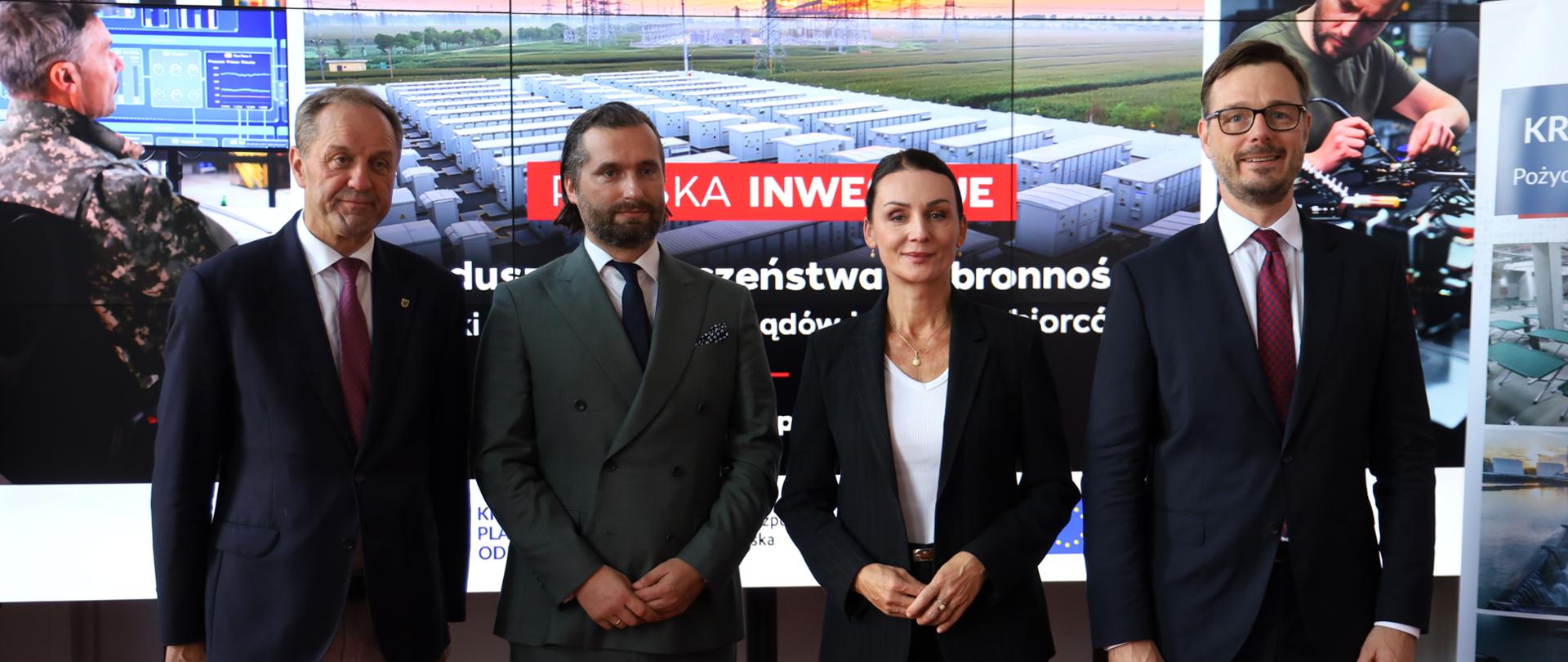 Cztery osoby w eleganckich strojach biznesowych pozują do zdjęcia na tle dużego ekranu z napisem „POLSKA INWESTUJE”.