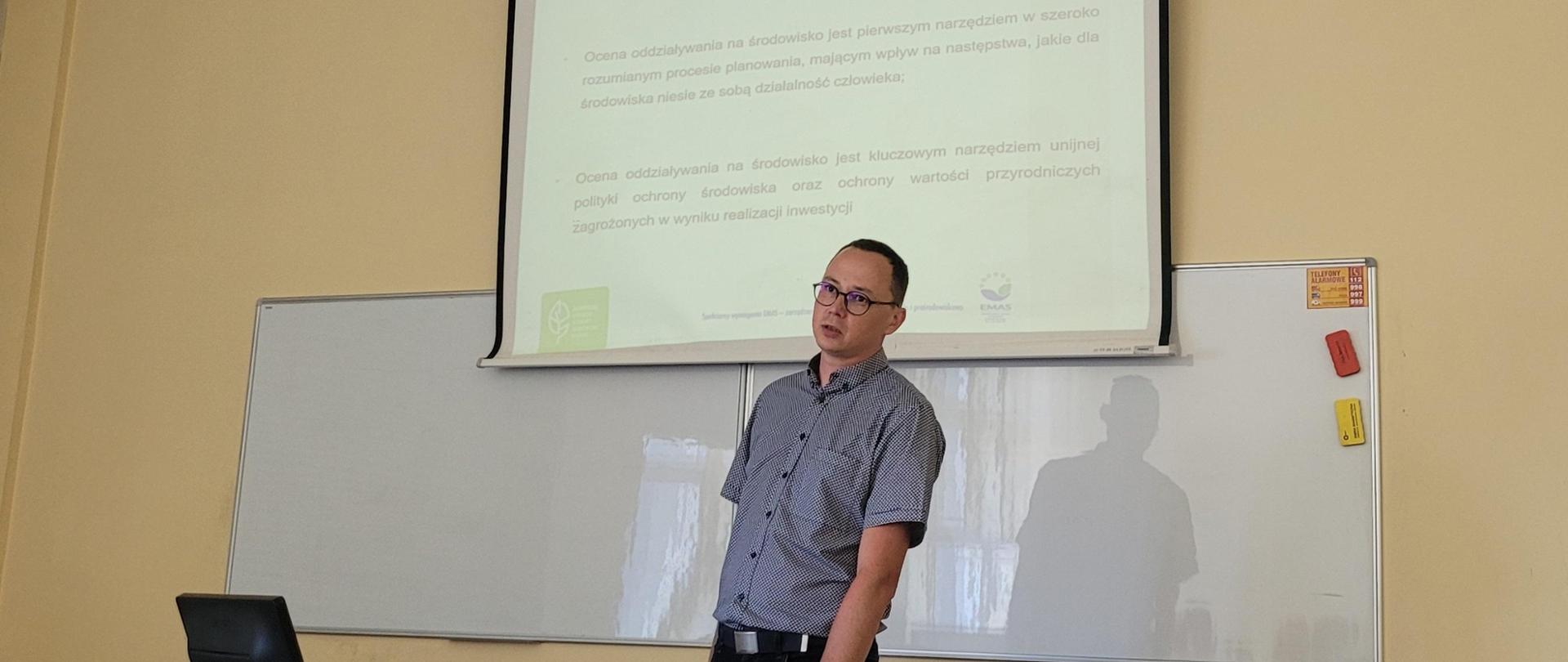 Mężczyzna stoi. Za nim beżowa ściana, u góry tablica z wyświetlonym tekstem, poniże pusta tablica.
Przed mężczyzną widoczny komputer. Mężczyzna patrzy przed siebie, mówi.