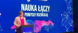 Za mównicą stoi wiceminister Zioło-Pużuk i mówi do mikrofonu, za nią wielki ekran z napisem na niebieskim tle Nauka łączy - pomysły rozwijają.