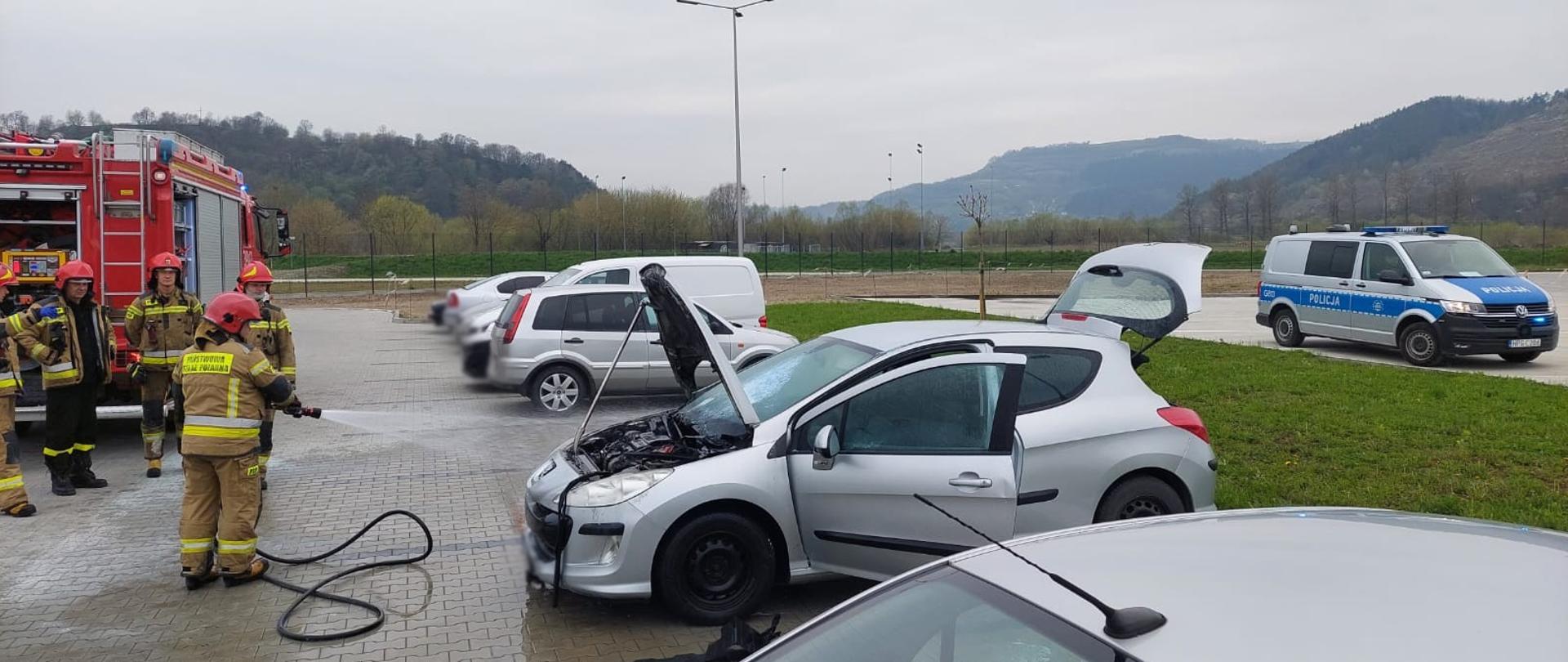 Strażacy gaszą pożar samochodu osobowego na parkingu.