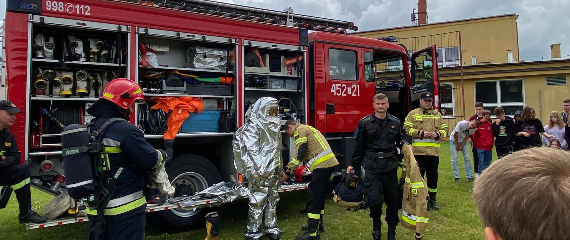 Na zdjęciu w porze dziennej widać na placu szkolnym na trawniku przed budynkiem szkoły strażaków pokazujących sprzęt pożarniczy. Strażacy stoją przodem do uczniów, tuż za nimi stoi samochód gaśniczy, pożarniczy.
