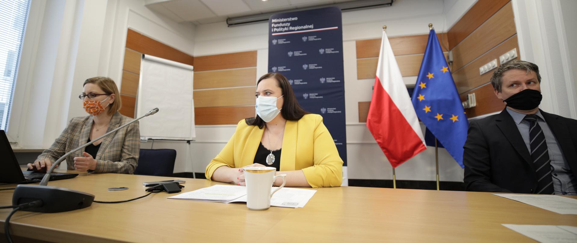 W sali konferencyjnej przy długim stole siedzą trzy osoby. Pośrodku wiceminister Małgorzata Jarosińska-Jedynak przed mikrofonem. Osoba z lewej strony obsługuje tablet. Z tyłu flagi PL i UE oraz rollup.