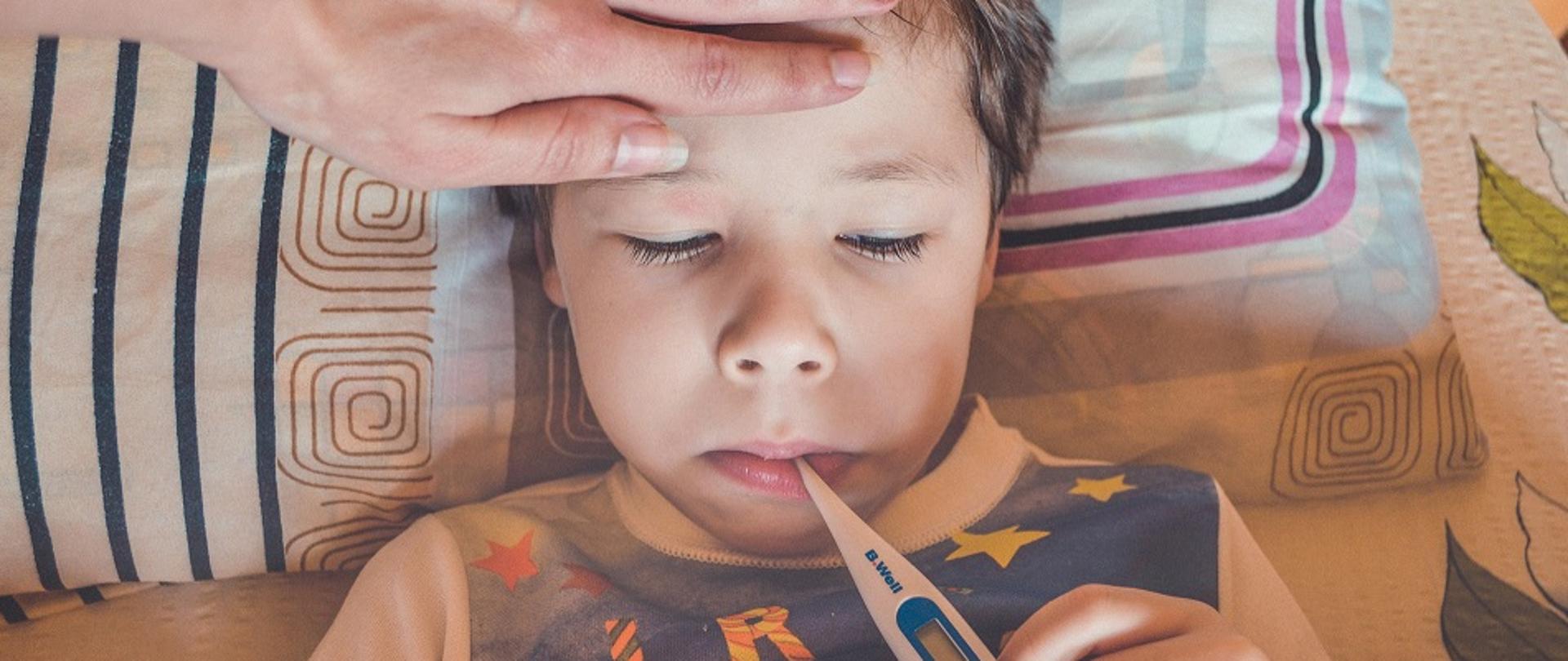 chłopiec leżący w łóżku z termometrem w ustach, ręka kobiety na czole, sprawdzająca także gorączkę