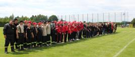 Zdjęcie grupowe młodzieżowych drużyn pożarniczych ustawionych w dwuszeregu na stadionie sportowym.