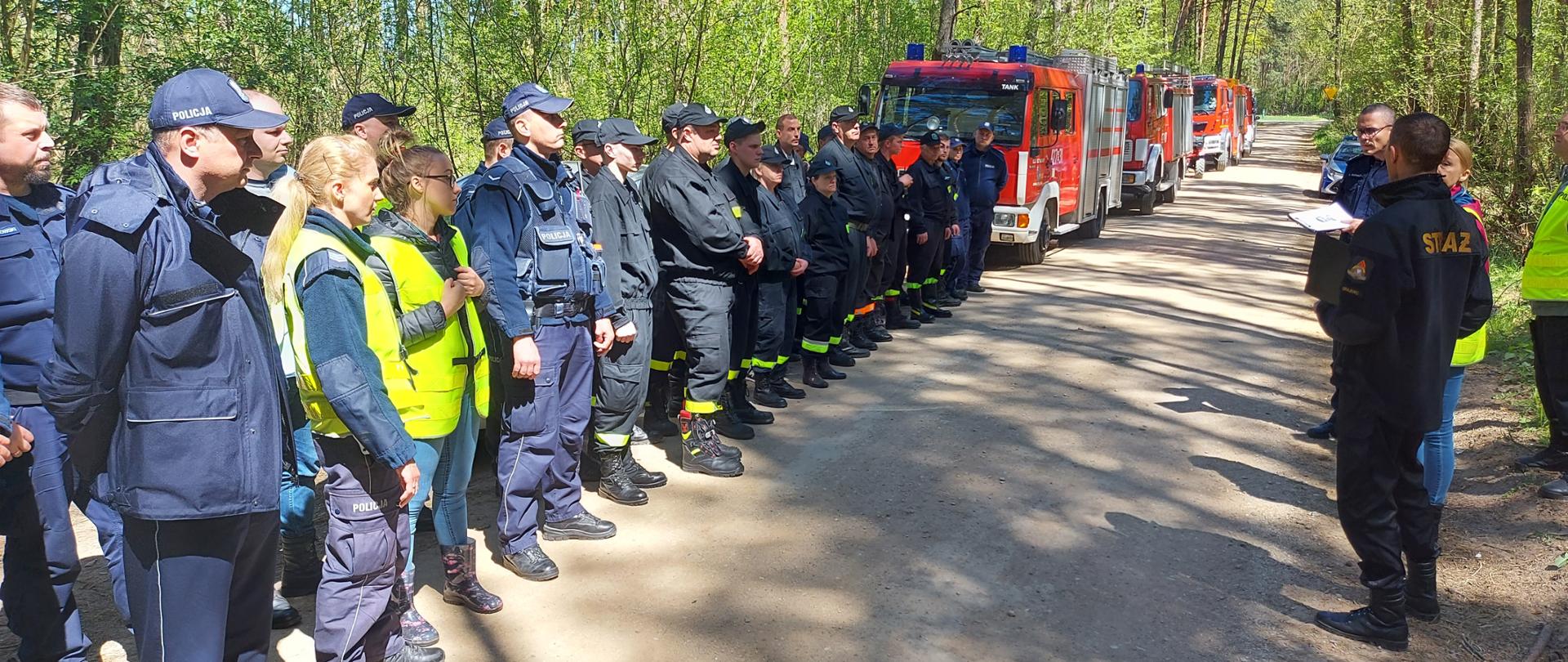 Zbiórka na drodze leśnej służb ratowniczych przed poszukiwaniami. W tle samochody straży pożarnej.