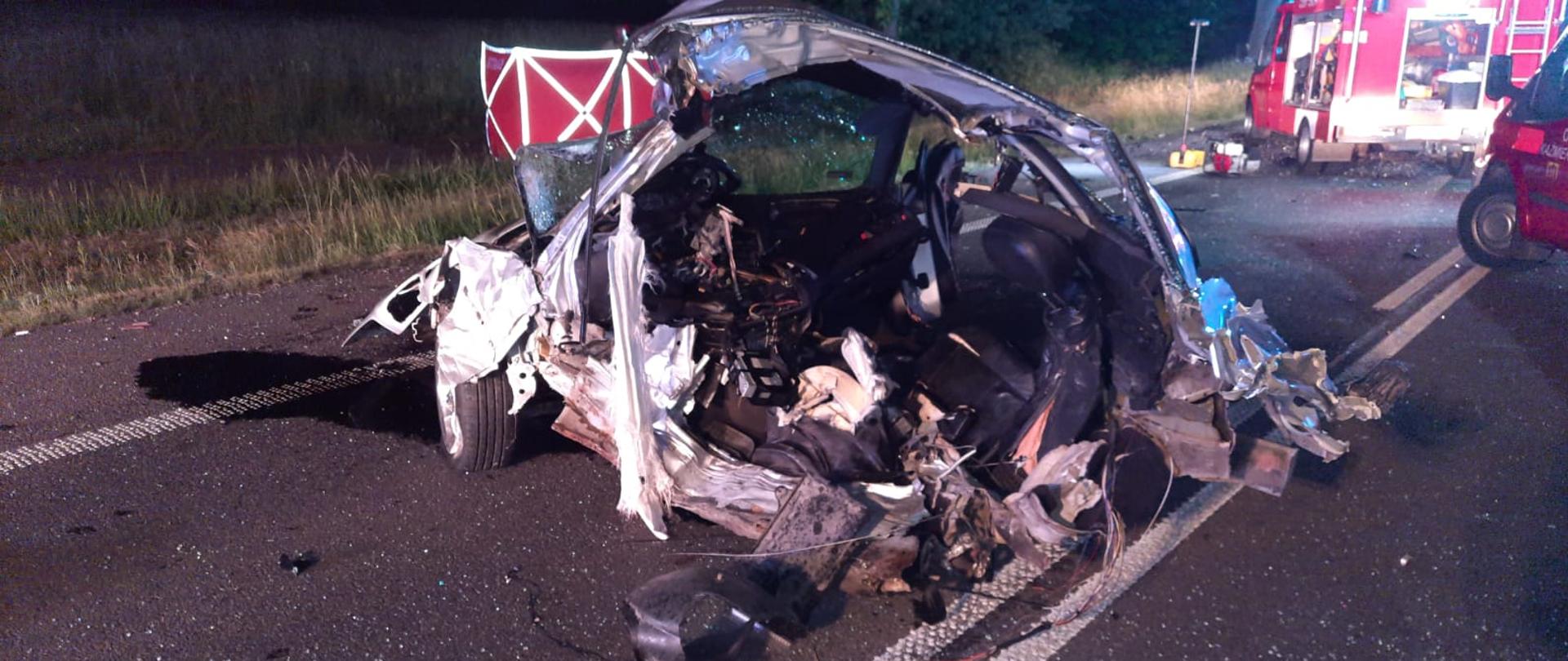 Widać część uszkodzonego pojazdu na drodze, widok lewego boku, po prawej samochód strażacki, jest noc