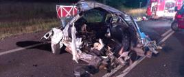 Widać część uszkodzonego pojazdu na drodze, widok lewego boku, po prawej samochód strażacki, jest noc