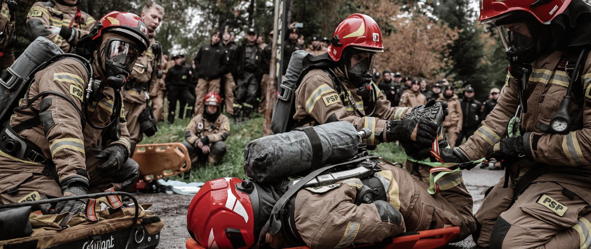 Strażacy w pełnym umundurowaniu ochronnym i maskach, z czerwonymi hełmami, pracują wokół osoby leżącej na pomarańczowych noszach ratunkowych. Osoba na noszach jest również w ubraniu strażackim i hełmie. Strażacy klęczą nad poszkodowanym, koncentrując się na wykonywanym zadaniu. Jeden z nich, po prawej stronie, trzyma ręce na poszkodowanym, podczas gdy inny, w centrum, wykonuje jakąś czynność związaną ze sprzętem. Trzeci strażak, po lewej stronie, klęczy obok torby ze sprzętem. W tle widać dużą grupę osób obserwujących sytuację oraz drzewa i leśne otoczenie.