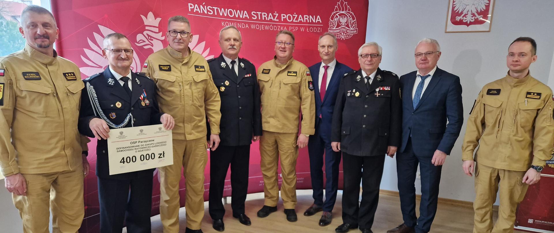 strażacy PSP, druhowie OSP, posłowie oraz przedstawiciele władz samorządowych pozują do zdjęcia na tle banneru komendy wojewódzkiej PSP w Łodzi