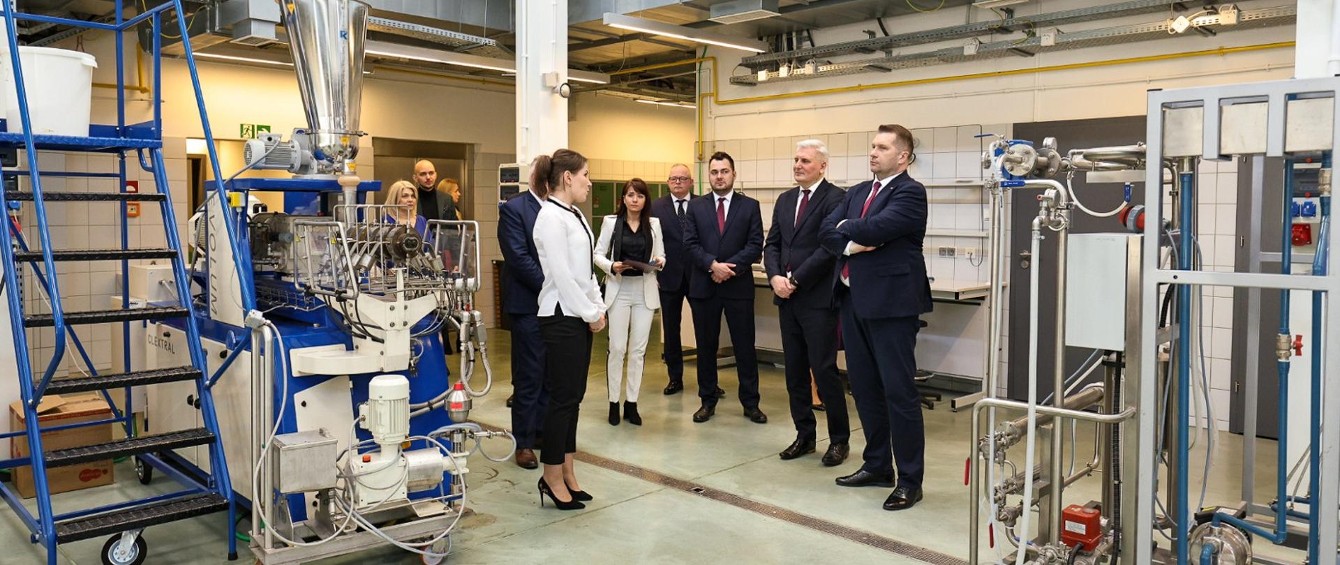 Minister Czarnek, kobieta w białej bluzce i kilkoro innych osób stoją na podłodze dużej hali, obok nich stoją różne skomplikowane urządzenia.