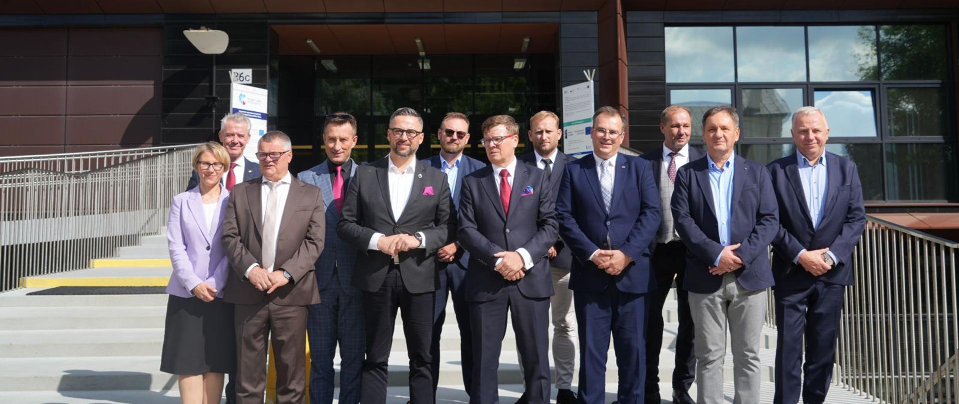 Przed schodami do wejścia do przeszklonego budynku stoi grupa kilkunastu osób, pośrodku grupy minister Kulasek.