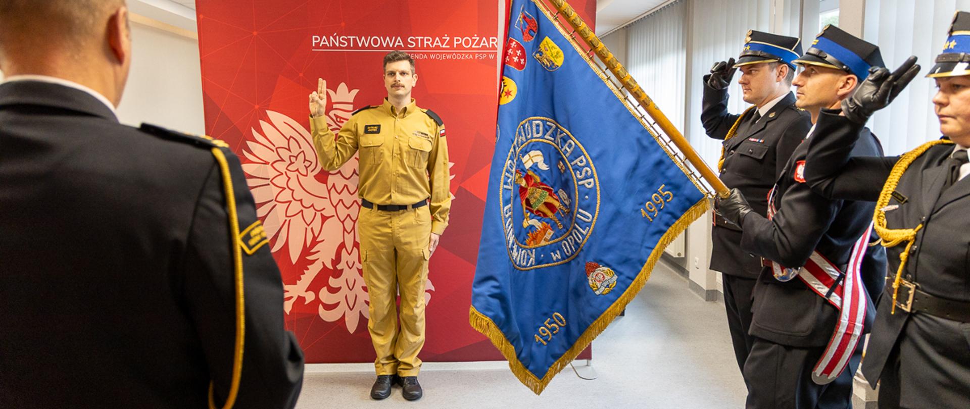 Osoba w jasnym, piaskowym mundurze PSP stoi przodem do pocztu sztandarowego i unosi prawą dłoń z trzema palcami w górze, wykonując gest ślubowania. Przed nim stoją trzej funkcjonariusze w ciemnych, galowych mundurach, jeden z nich trzyma niebieski sztandar PSP z haftowanymi symbolami i datami. W tle widoczna jest czerwona ściana z dużym godłem Polski oraz napisem „Państwowa Straż Pożarna Komenda Wojewódzka PSP w Opolu”. Scena przedstawia uroczysty moment wypowiedzenia roty ślubowania przez str. Mateusza Andresa.
