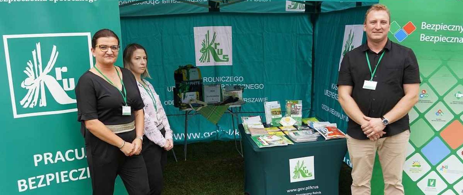 Zielony namiot promocyjny KRUS wyposażony w roll-upy i materiały promocyjne. Przed namiotem dwie kobiety i mężczyzna – pracownicy KRUS, uśmiechają się do zdjęcia.