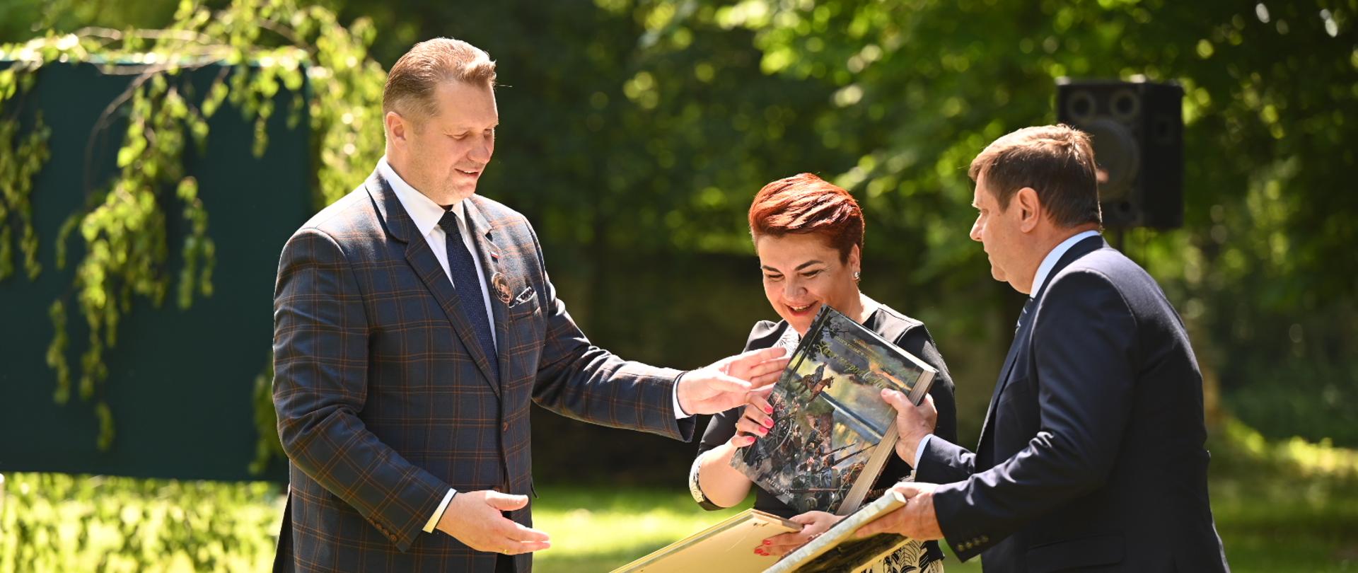 Minister podaje dyplom w ramce mężczyźnie w garniturze, obok stoi kobieta w sukience w kwiatki.