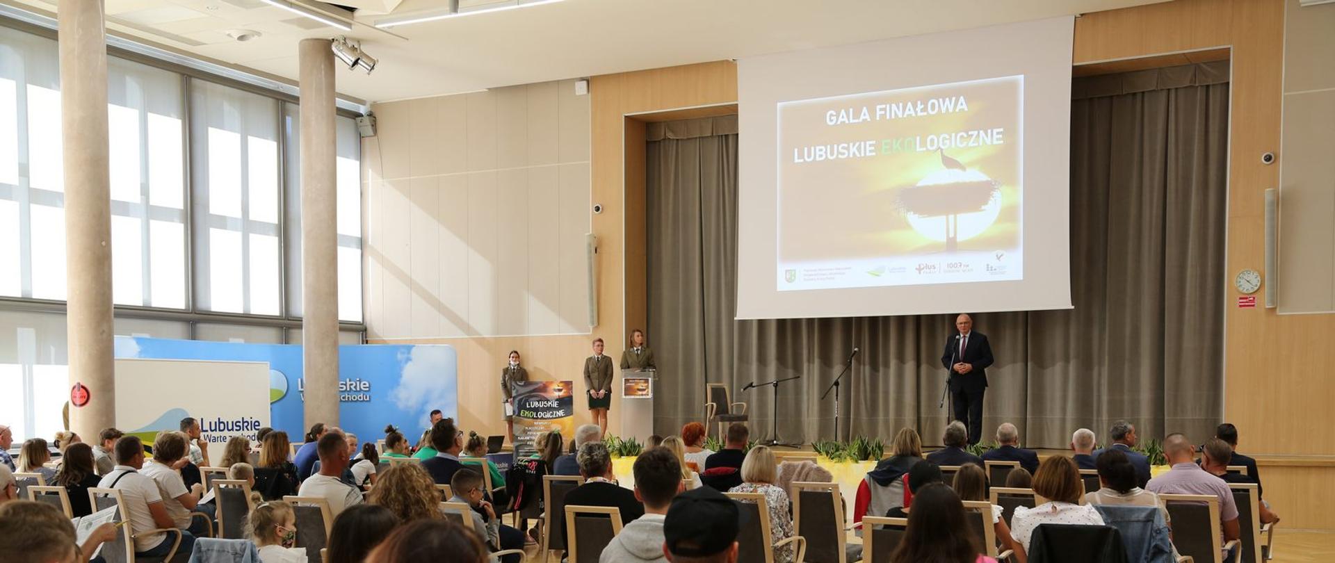 W dużej sali na krzesłach odwrócone plecami siedzi wiele osób. W tle na scenie stoi mężczyzna, mówiący do mikrofonu. Po lewej stornie stoją trzy kobiety.