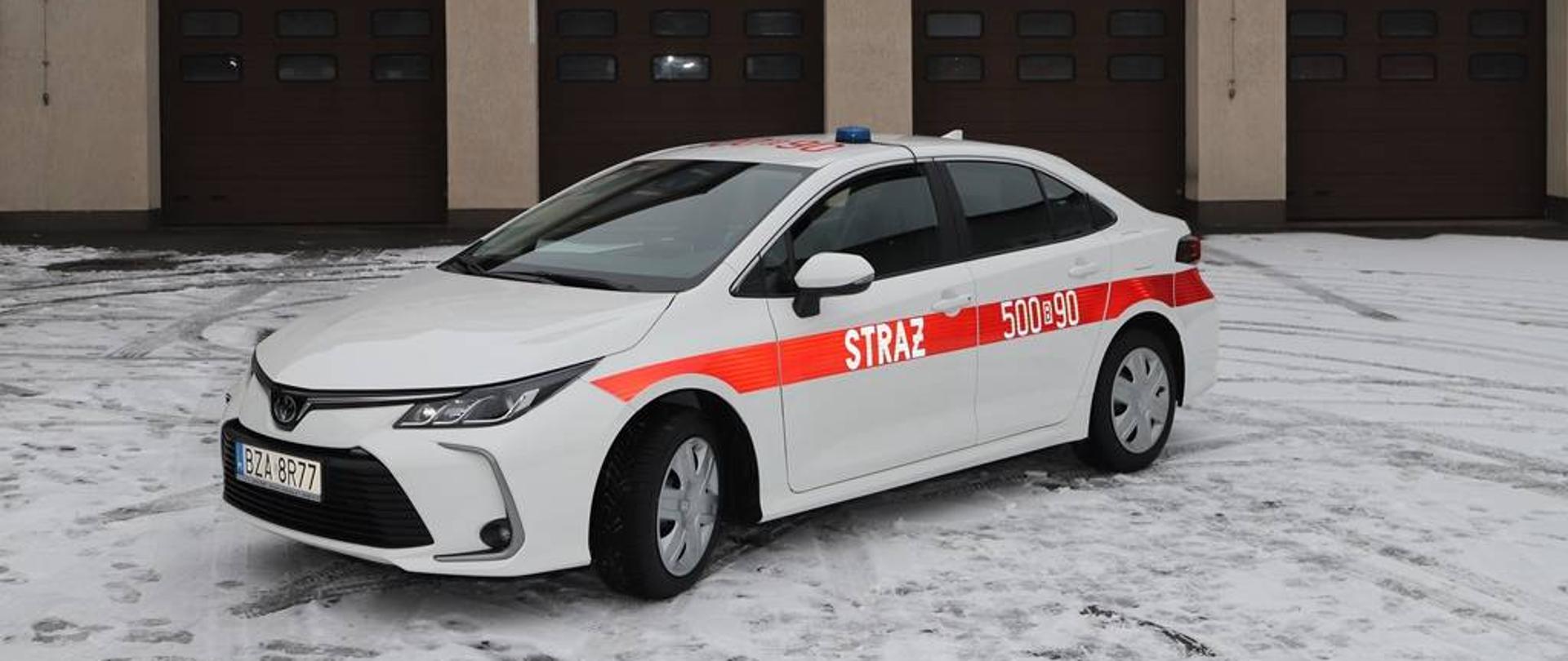 Zdjęcie przedstawia nowy samochód operacyjny Komendy Powiatowej Państwowej Straży Pożarnej w Zambrowie. Biała Toyota Corolla z czerwonym pasem przebiegającym wzdłuż boku samochodu na którym widnieje napis "STRAŻ" oraz numer operacyjny 500B90. W tle widać budynek Komendy Powiatowej Państwowej Straży Pożarnej w Zambrowie.