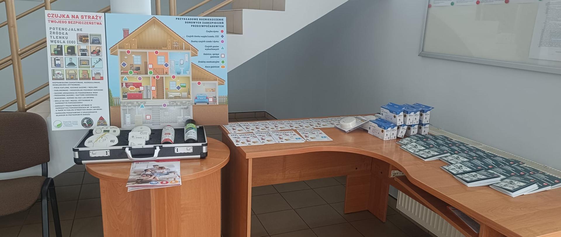 Widok na przygotowane stoisko profilaktyczne w holu budynku. Na okrągłym stoliku stoi makieta domu oraz otwarta walizka z różnymi rodzajami czujek (czadu, dymu, gazu) i sprayami gaśniczymi. Na sąsiednim biurku ułożone są ulotki i broszury informacyjne.