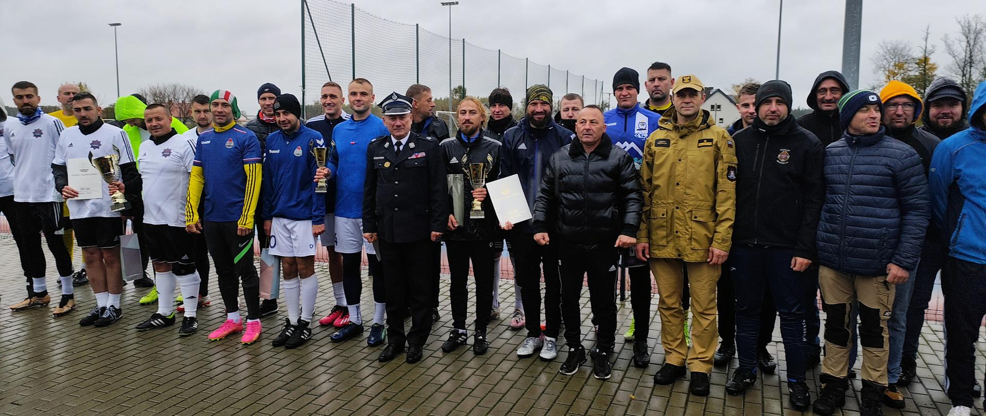 Grupa mężczyzn, z których wielu jest ubranych w stroje sportowe lub ciepłe kurtki, stoi na mokrym bruku na zewnątrz boiska sportowego. Kilku z nich trzyma puchary i dyplomy, jest to ceremonia wręczenia nagród p , meczu piłki nożnej, a na pierwszym planie wyróżniają się mężczyźni w mundurach.