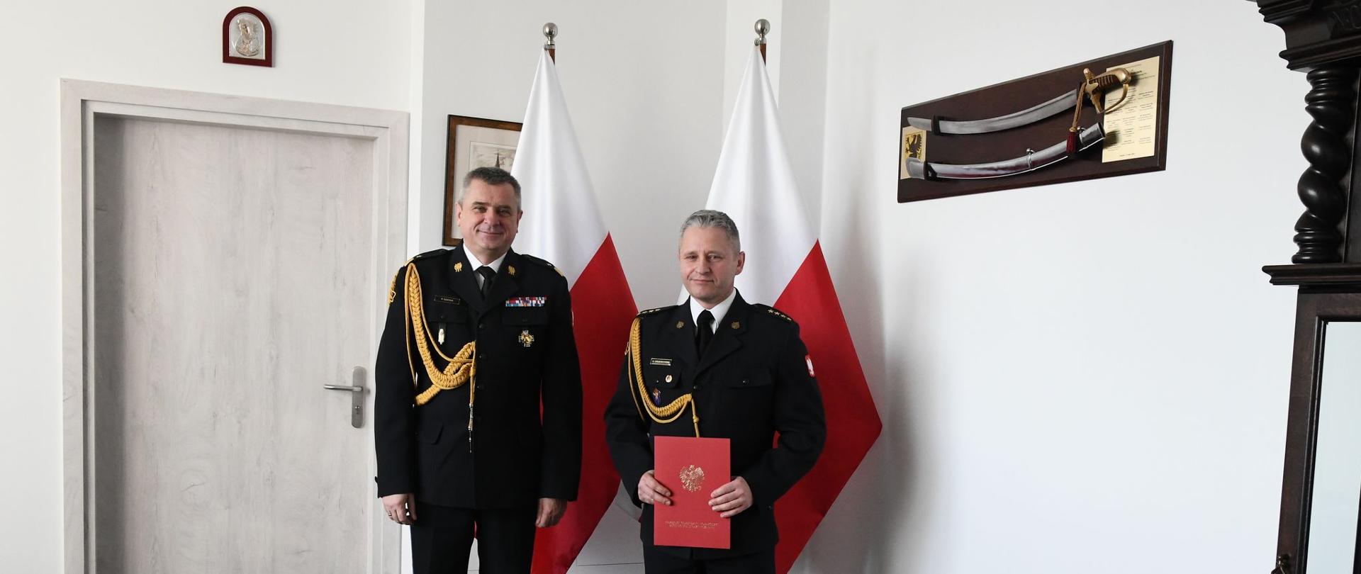 Funkcjonariusze Państwowej Straży Pożarnej stoją obok siebie jedne z nich trzyma czerwoną teczkę za nimi znajdują się flagi Polski.